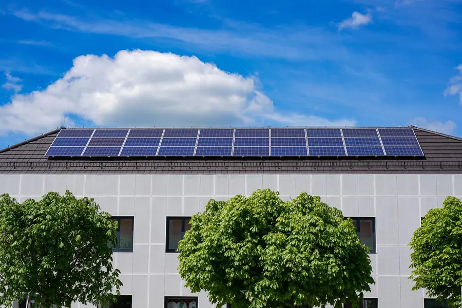 Office building with a row of solar panels along its roof edge, set among trees under a partly cloudy sky