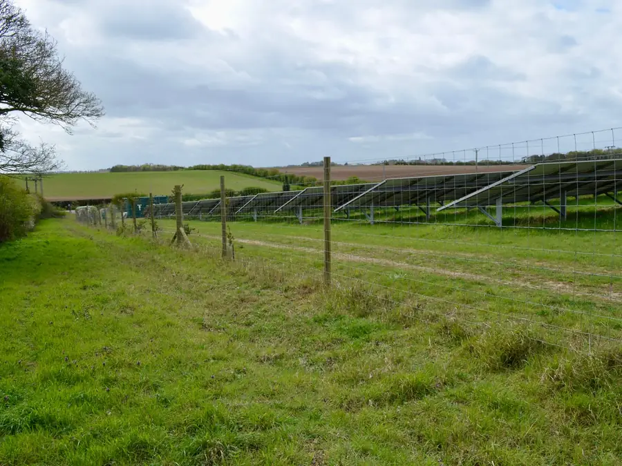 Fenced ground-mounted solar field running behind a hedgerow and post-and-wire fence on farmland